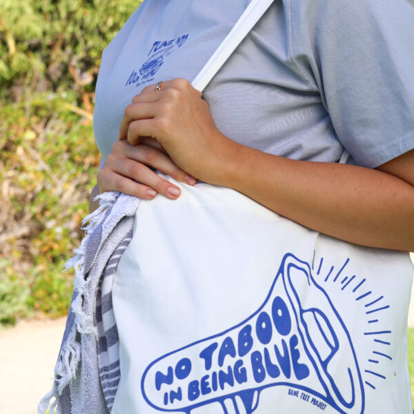 Natural cotton tote bag with bold blue text reading ‘No Taboo in Being Blue’, held by a person standing outdoors, promoting mental health awareness