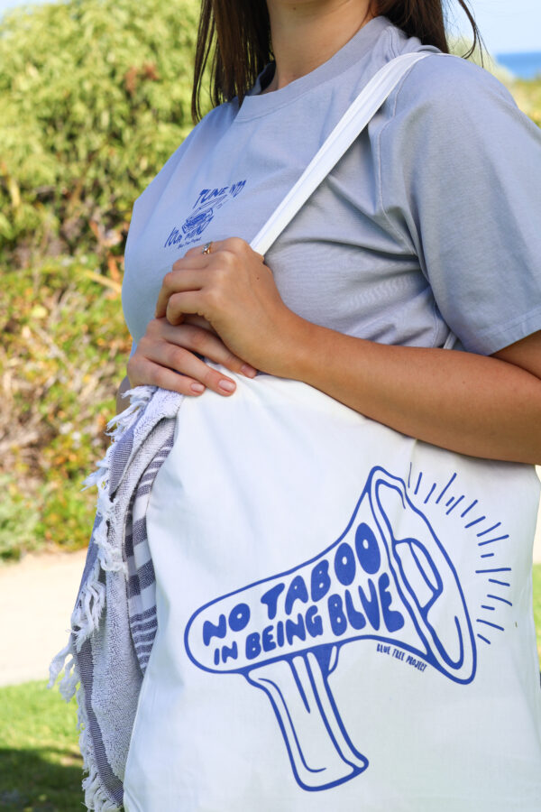 Natural cotton tote bag with bold blue text reading ‘No Taboo in Being Blue’, held by a person standing outdoors, promoting mental health awareness